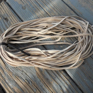 May include: A bundle of light brown leather cord, approximately 3 metres long, is coiled on a wooden surface. The cord is made of thin strips of leather that are woven together.