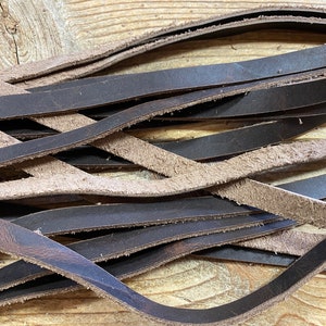 May include: A pile of brown leather strips, some with a suede finish, arranged in a criss-cross pattern on a wooden surface.