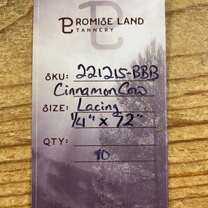 May include: A white card with black text. The text reads "PROMISE LAND TANNERY" in a stylized font. Below the text is a product description: "SKU: 221215-BBB Cinnamon Cow SIZE: Lacing 1/4" x 72" QTY: 10".