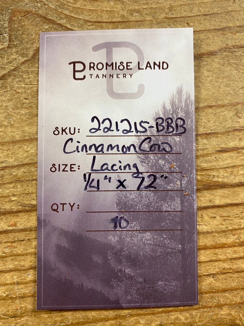 May include: A white card with black text. The text reads "PROMISE LAND TANNERY" in a stylized font. Below the text is a product description: "SKU: 221215-BBB Cinnamon Cow SIZE: Lacing 1/4" x 72" QTY: 10".