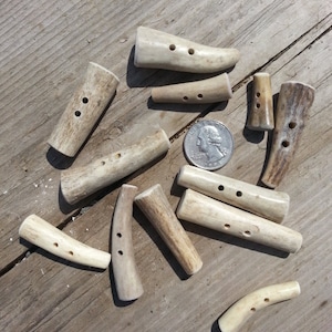 May include: A collection of 10 natural antler buttons with two holes each. The buttons are various shapes and sizes, with some being curved and others straight. The buttons are a light brown color.