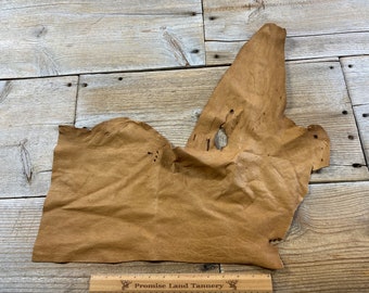 May include: A piece of brown leather with a rough texture. The leather is folded and has a few holes in it. It is sitting on a wooden surface.