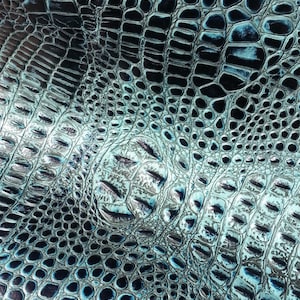 May include: Close-up of a teal blue and black crocodile embossed leather fabric. The fabric has a textured, scaly pattern.