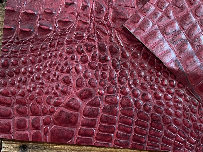 May include: Close-up of a burgundy faux crocodile leather fabric with a textured, embossed pattern.