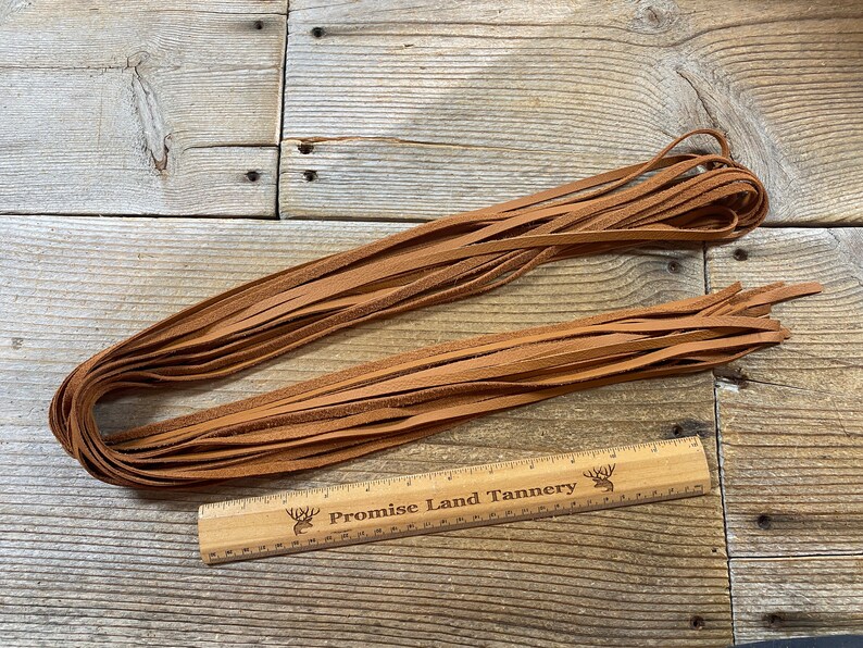May include: A bundle of brown leather strips, approximately 12 inches long, laid out on a wooden surface. A ruler with the text "Promise Land Tannery" is placed next to the leather strips.