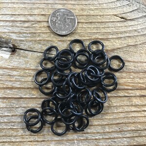 May include: A pile of black jump rings, approximately the size of a US quarter, on a wooden surface.