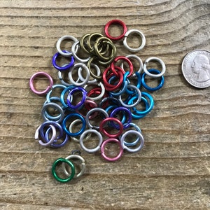 May include: A collection of colorful metal jump rings in various sizes, including silver, gold, red, blue, green, and purple. The rings are arranged in a pile on a wooden surface. A US quarter is included for scale.