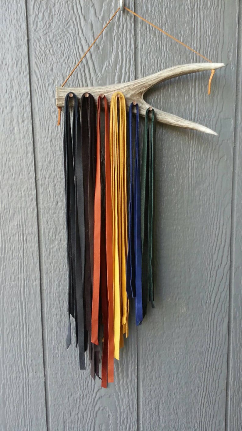 May include: A set of leather cords hanging from a wooden antler rack. The cords are in various colors, including black, brown, orange, yellow, and blue.