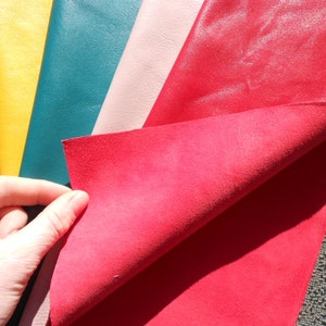 May include: A close-up of five different colored leather swatches. The swatches are arranged in a fan shape, with the top swatch folded over to reveal the underside. The colors of the swatches are yellow, teal, pink, red, and a darker red.