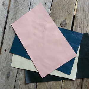 May include: Four pieces of leather in different colors: pink, cream, teal, and dark green. The leather is laid out on a wooden surface.