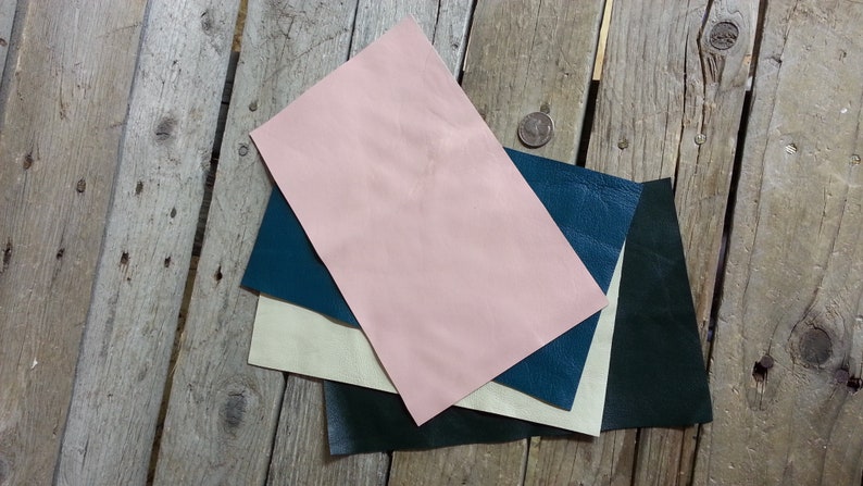 May include: Four pieces of leather in different colors: pink, cream, teal, and dark green. The leather is laid out on a wooden surface.