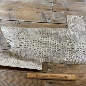 May include: Off-white alligator-textured leather pieces. The leather has a detailed pattern of scales and ridges, with a central oval design. A wooden ruler with text is at the bottom. The leather is on a wooden surface.