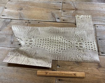 May include: Off-white alligator-textured leather pieces. The leather has a detailed pattern of scales and ridges, with a central oval design. A wooden ruler with text is at the bottom. The leather is on a wooden surface.