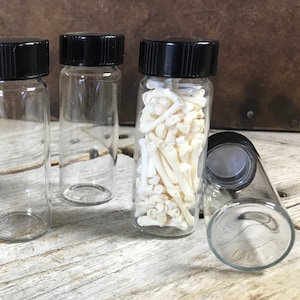 May include: Small glass vials with black caps, some empty and some filled with small, off-white bones. The vials are arranged on a weathered, light-colored wooden surface, with a rustic, brown background.