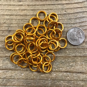 May include: A pile of gold-colored metal jump rings, approximately 1/2 inch in diameter, next to a US quarter for size comparison.