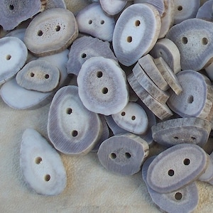 May include: A collection of natural antler buttons in various sizes and shapes. The buttons are a light gray color with a smooth, polished finish. They have two or three holes for sewing.
