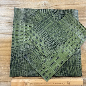May include: Three pieces of olive green faux leather with a crocodile skin texture. The leather is arranged on a wooden surface. A ruler with the text "Promise Land Tannery" is at the bottom.