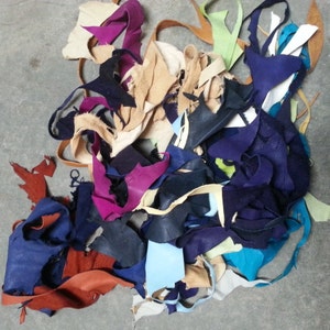 May include: A pile of leather scraps in various colors, including blue, purple, tan, and green, are scattered on a gray surface. The leather pieces vary in size and shape, suggesting they are remnants from a larger project.