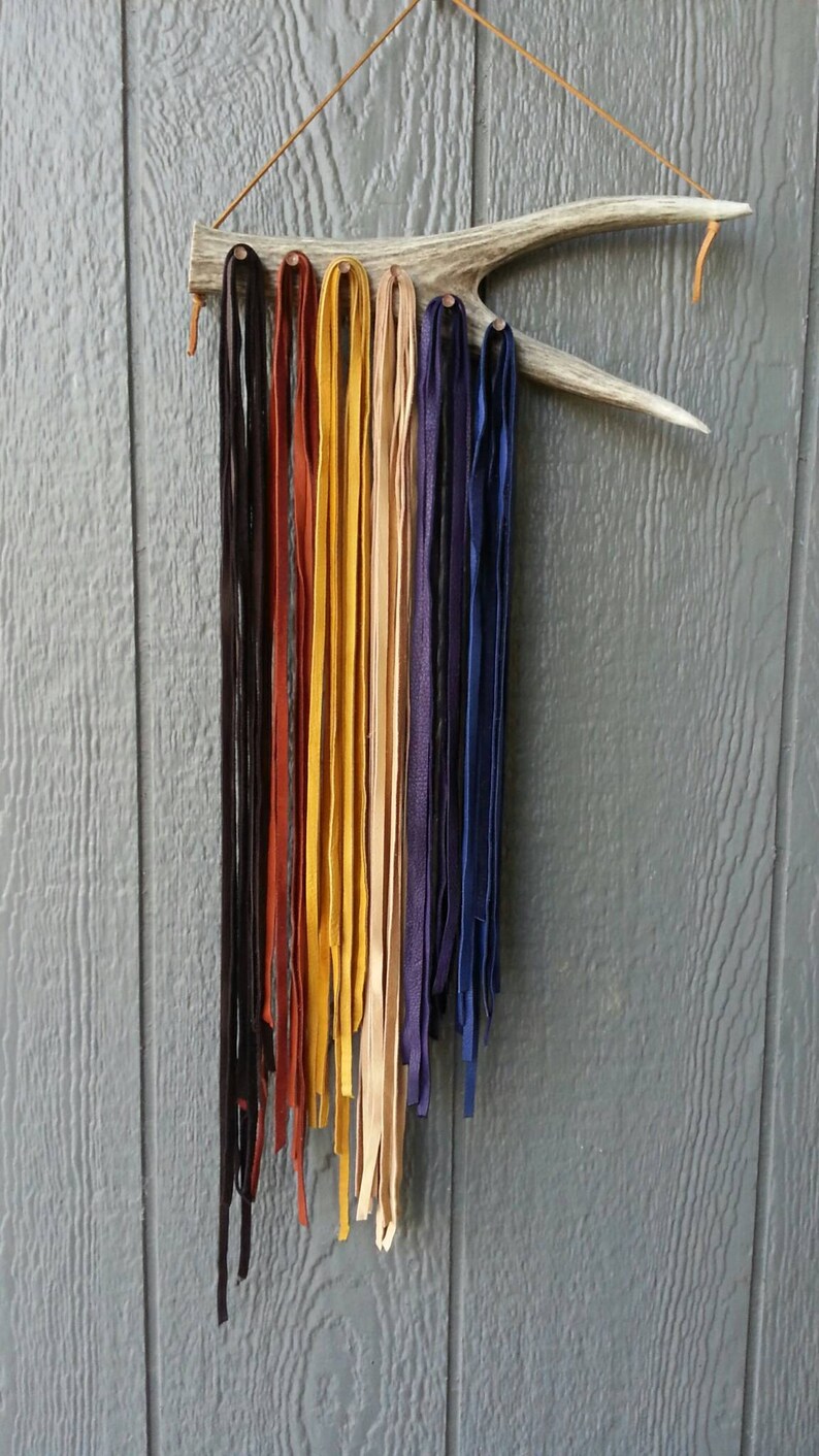 K&ouml;nnte beinhalten: Ein Satz Lederstreifen in verschiedenen Farben, darunter Schwarz, Braun, Gelb, Beige und Lila, die an einem Holzgeweih h&auml;ngen.