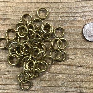 May include: A pile of gold jump rings, approximately the size of a US quarter, on a wooden surface.