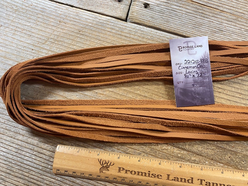 May include: A bundle of 10 cinnamon colored leather lacing strips, each measuring 1/4 inch wide by 72 inches long. The strips are stacked neatly on a wooden surface. The label reads "SKU: 221215-BBB Cinnamon Co Lacing 1/4" x 72" QTY: 10" and is from Promise Land Tannery.