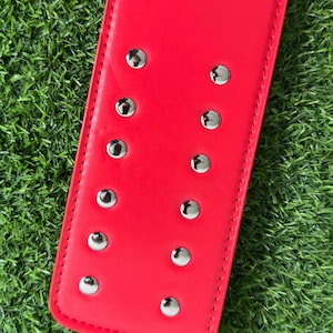 May include: Close-up of a red leather belt with silver studs. The belt is on a green grassy surface.