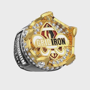 Puede incluir: Un anillo dorado con una cara ornamentada. La cara presenta la palabra "GRIDIRON", un diseño de flor de lis y numerosas piedras preciosas transparentes. La banda del anillo es plateada con la palabra "GRIDIRON".