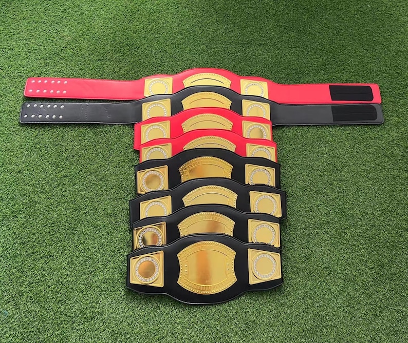 May include: A stack of black and red championship belts with gold accents. Each belt has a large gold plate in the center with a smaller gold circle on each side. The belts are made of leather and have a buckle closure.