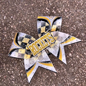 May include: A large cheerleading bow with a gold glitter center that reads "ROCKETS". The bow features a black, white, and yellow checkered pattern with rhinestone accents. The bow is made of ribbon and is designed for cheerleading.