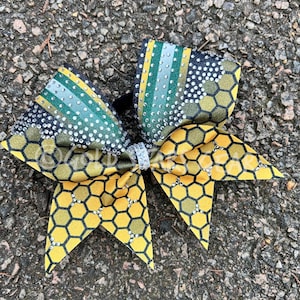May include: A large, decorative hair bow featuring a honeycomb pattern in yellow and black, accented with silver rhinestones. The bow also has stripes of green and silver, and is set against a textured surface.