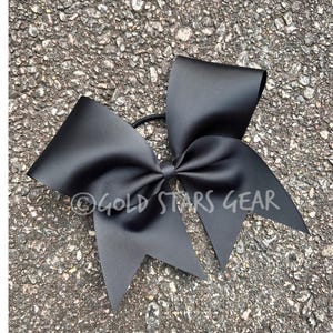 May include: A large, black hair bow with a ribbed texture, secured with a black elastic band. The bow is centered and has a classic, symmetrical design. The text "GOLD STARS GEAR" is printed on the bow.