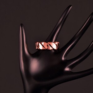 Pode incluir: Um anel de cobre com um design torcido. O anel é exibido em um modelo de mão preto.