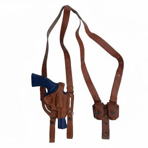 May include: A brown leather shoulder holster with a blue handgun. The holster includes a double magazine pouch and adjustable straps. The background is plain white.