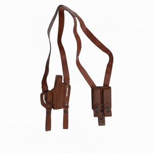 May include: A brown leather shoulder holster with a holster for a handgun and a double magazine pouch. The straps are adjustable and have metal hardware. The background is white.