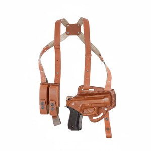 May include: A brown leather shoulder holster with a matching magazine pouch. The holster is designed to hold a handgun, and the straps are adjustable. The leather has a smooth finish and is stitched together with a lighter-colored thread.