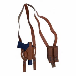May include: A brown leather shoulder holster with a blue handgun and a double magazine pouch. The holster has adjustable straps and is designed to be worn across the chest and back. The background is plain white.