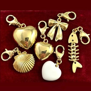 May include: Six gold-tone charm pendants with lobster claw clasps. The charms include a heart, a seashell, a bow, a fish bone, and two hearts.