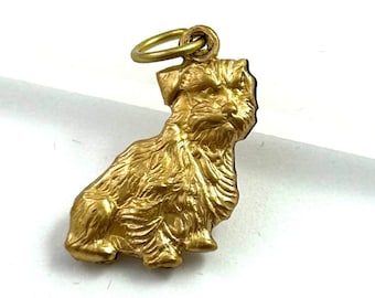 Puede incluir: Un amuleto de color dorado de un perro sentado con un diseño de pelaje texturizado y detallado. El amuleto tiene un bucle en la parte superior para sujetarlo a un collar o pulsera.