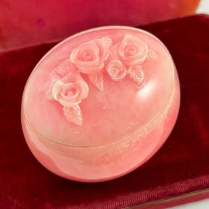 May include: A pink oval-shaped trinket box with a carved rose design on the lid.