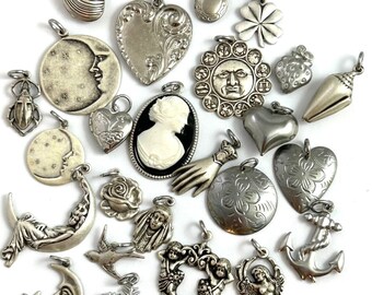 May include: A collection of 20 silver charm pendants featuring various designs, including a crescent moon, a heart, a rose, a snowflake, an anchor, a bird, a hand, a seashell, a four-leaf clover, a ladybug, and a cameo.