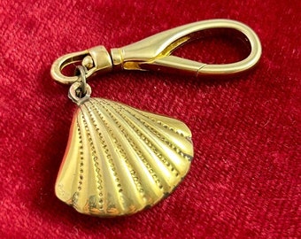 May include: A gold-colored seashell charm attached to a gold-colored key chain. The seashell has a detailed, textured design.