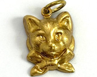May include: A gold-colored charm in the shape of a cat's head with a bow tie. The charm has a loop at the top for attaching to a necklace or bracelet.