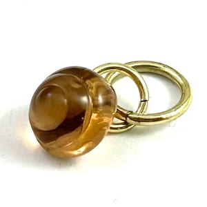 May include: A translucent amber-colored glass bead with a smooth, rounded shape attached to two interlocking gold-toned metal jump rings. The bead features a swirling internal pattern and a glossy finish, connected to the circular metal rings.