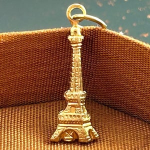 May include: A gold-colored Eiffel Tower charm pendant. The charm is detailed with intricate carvings and has a loop at the top for attaching to a necklace or bracelet.