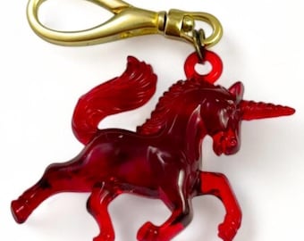 Puede incluir: Un llavero de plástico rojo en forma de unicornio con un cierre de metal dorado. El unicornio está en una pose de carrera con su cuerno apuntando hacia adelante.