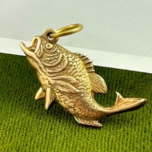 May include: A gold-toned fish charm pendant. The fish is depicted with its mouth open and its fins spread out. The charm has a loop at the top for attaching to a chain or bracelet.