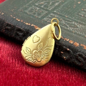 May include: A gold teardrop-shaped locket with a floral design and a small heart engraved on the front. The locket has a hinged opening and a loop for attaching a chain.