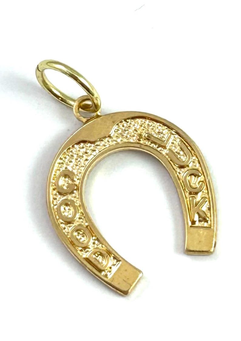May include: A gold-colored horseshoe pendant with the word "LUCK" inscribed. The pendant has a textured surface and a small loop for a chain. The horseshoe shape is a classic symbol of good fortune.