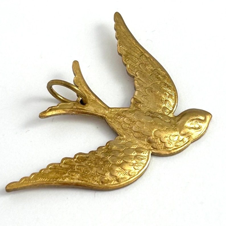 May include: A gold-toned metal pendant in the shape of a stylized bird with detailed wings and a loop for hanging.