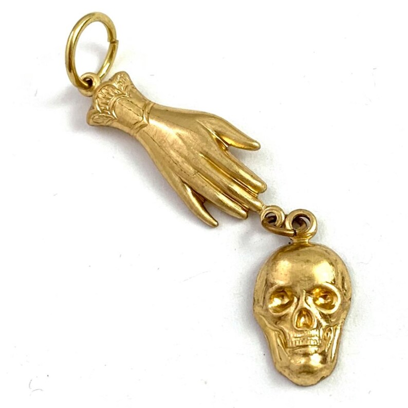 Op de afbeelding: Een gouden bedelhanger met een gedetailleerde hand die naar een schedel reikt. De hand is gestyled met een manchet aan de pols en de schedel heeft een gedetailleerd gezicht.